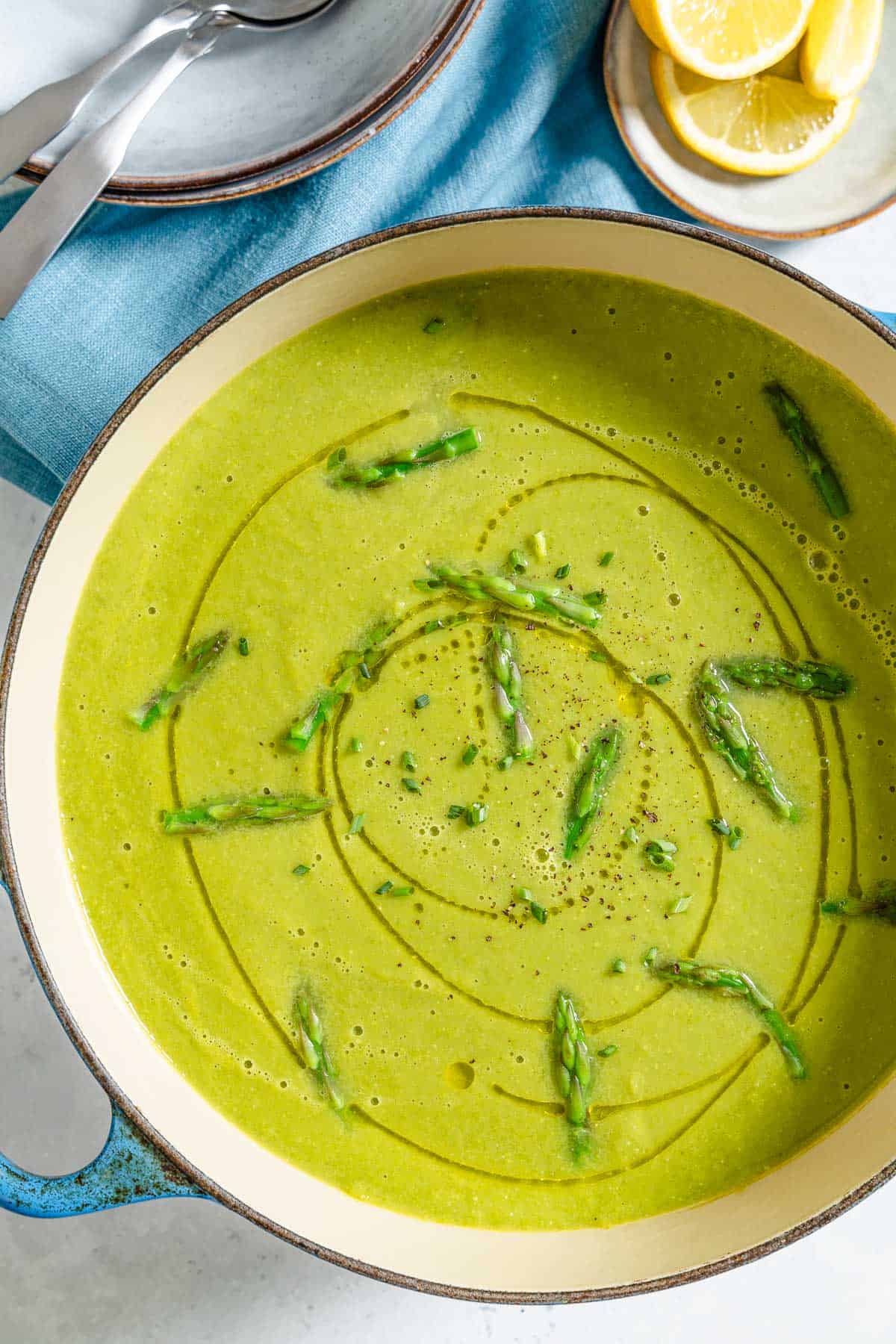 An overhead photo of a pot of asparagus soup. Next to this is a small plate with lemon wedges, and a stack of 2 plates with 2 spoons on top.