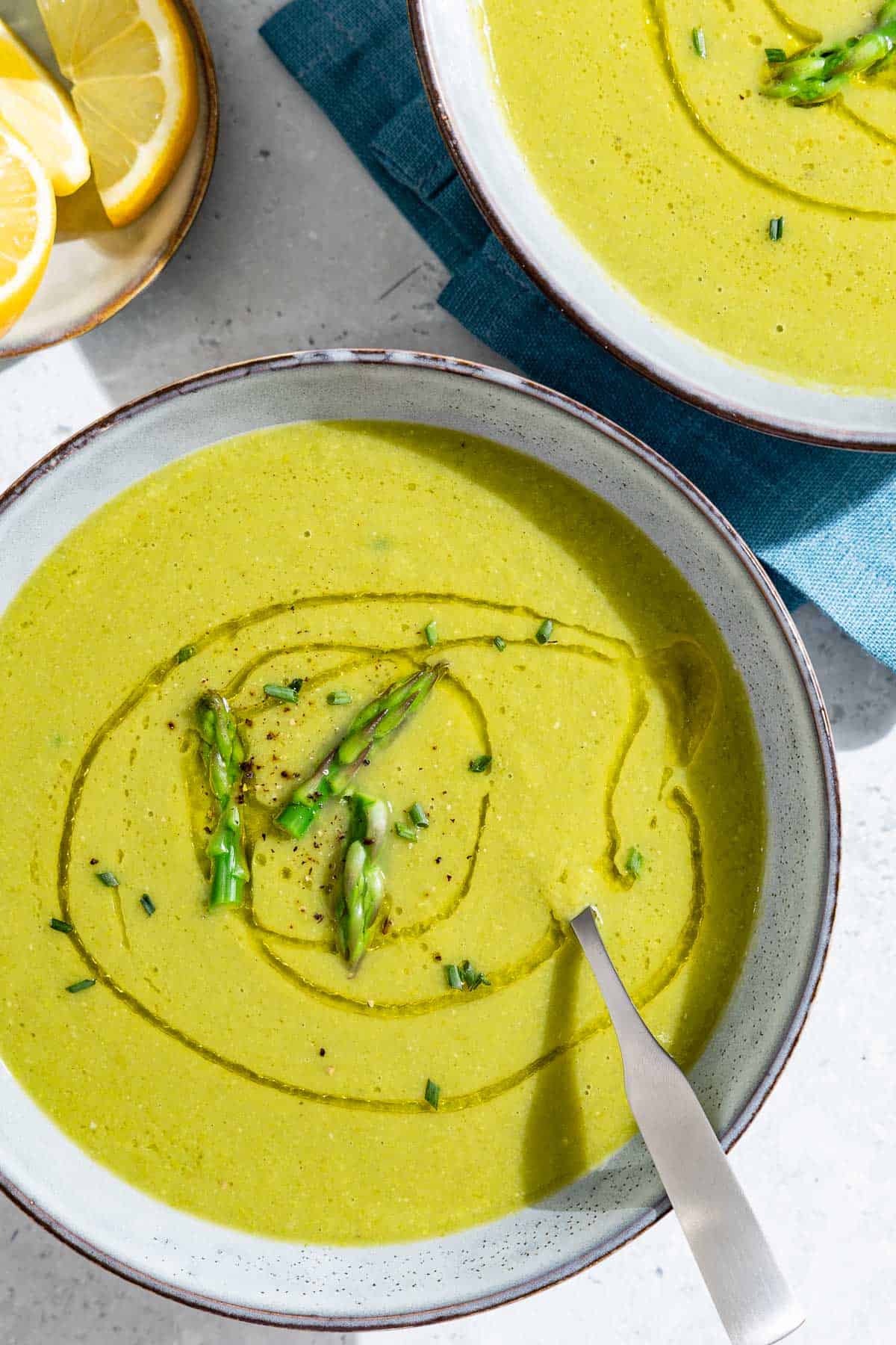 An overhead photo of two bowls of asparagus soup, one with a spoon. Next to these is a small plate of lemon wedges.