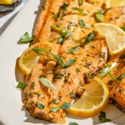 An overhead photo of baked trout garnished with parsley and green onions on a serving platter with lemon wedges.