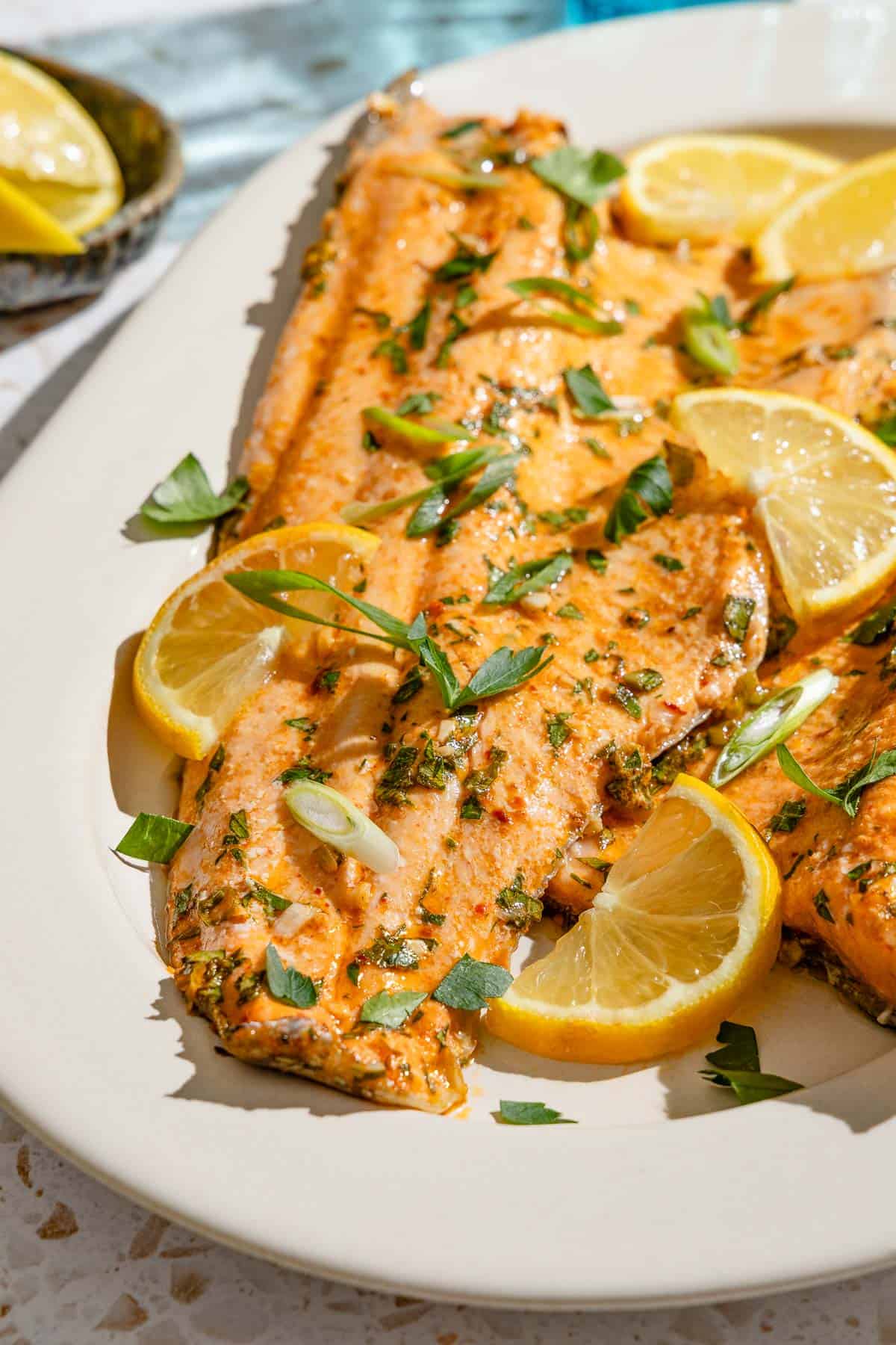 An overhead photo of baked trout garnished with parsley and green onions on a serving platter with lemon wedges.