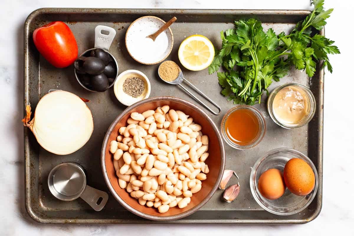 The ingredients for cannellini bean salad including cannellini beans, onion, tomato, parsley, black olives, salt, pepper, garlic, lemon, cumin, tahini, apple cider vinegar, water, eggs, and olive oil.
