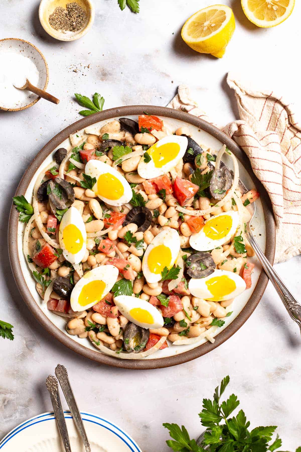 An overhead photo of cannellini bean salad on a serving platter with a fork. Next to this is a stack of plates with forks, small bowls of salt and pepper, 2 lemon halves, a cloth napkin, and a bunch of parsley.