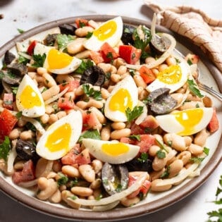 A close up of cannellini bean salad on a serving platter with a fork. Next to this is a bowl of salt and a cloth napkin.