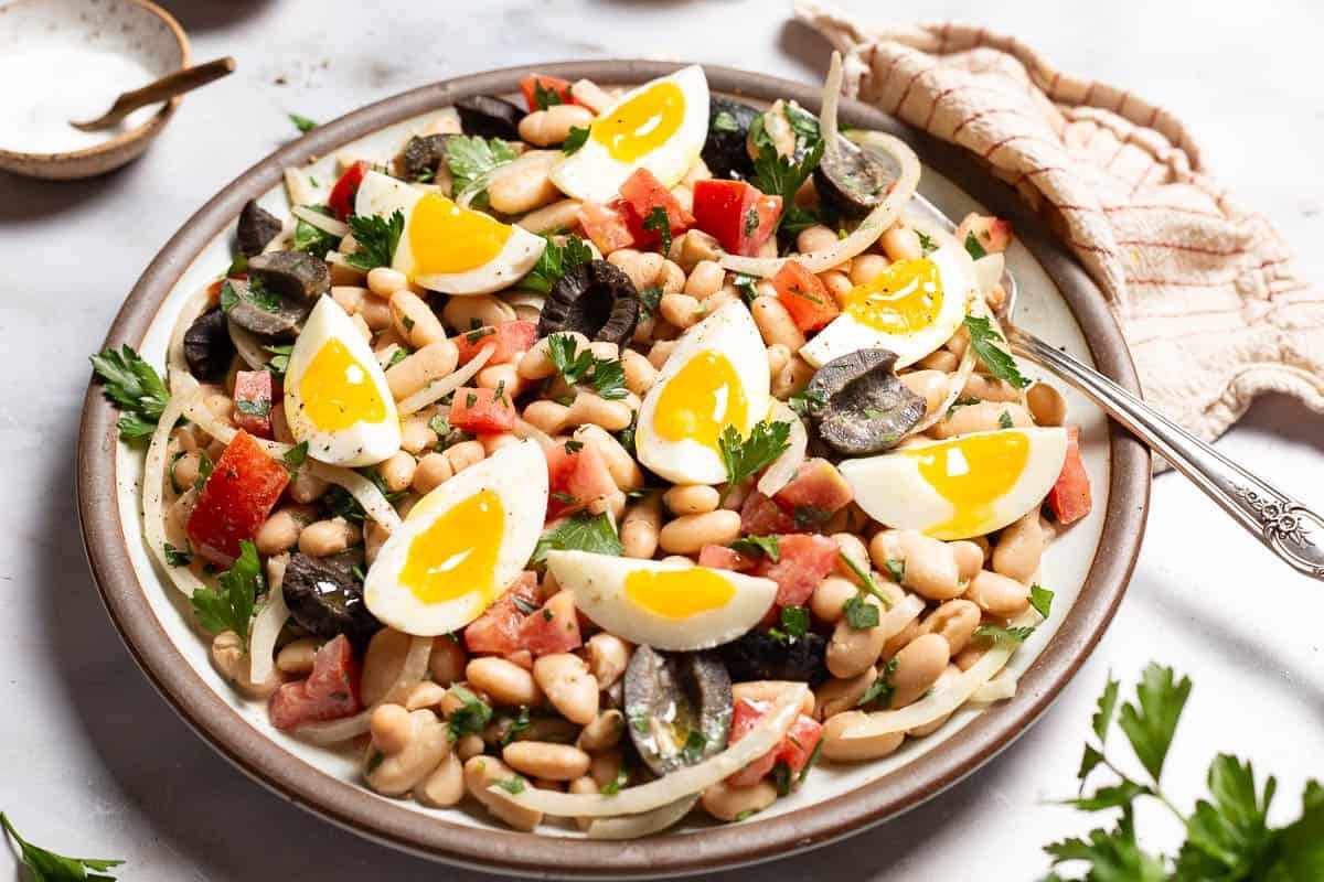 A close up of cannellini bean salad on a serving platter with a fork. Next to this is a bowl of salt and a cloth napkin.