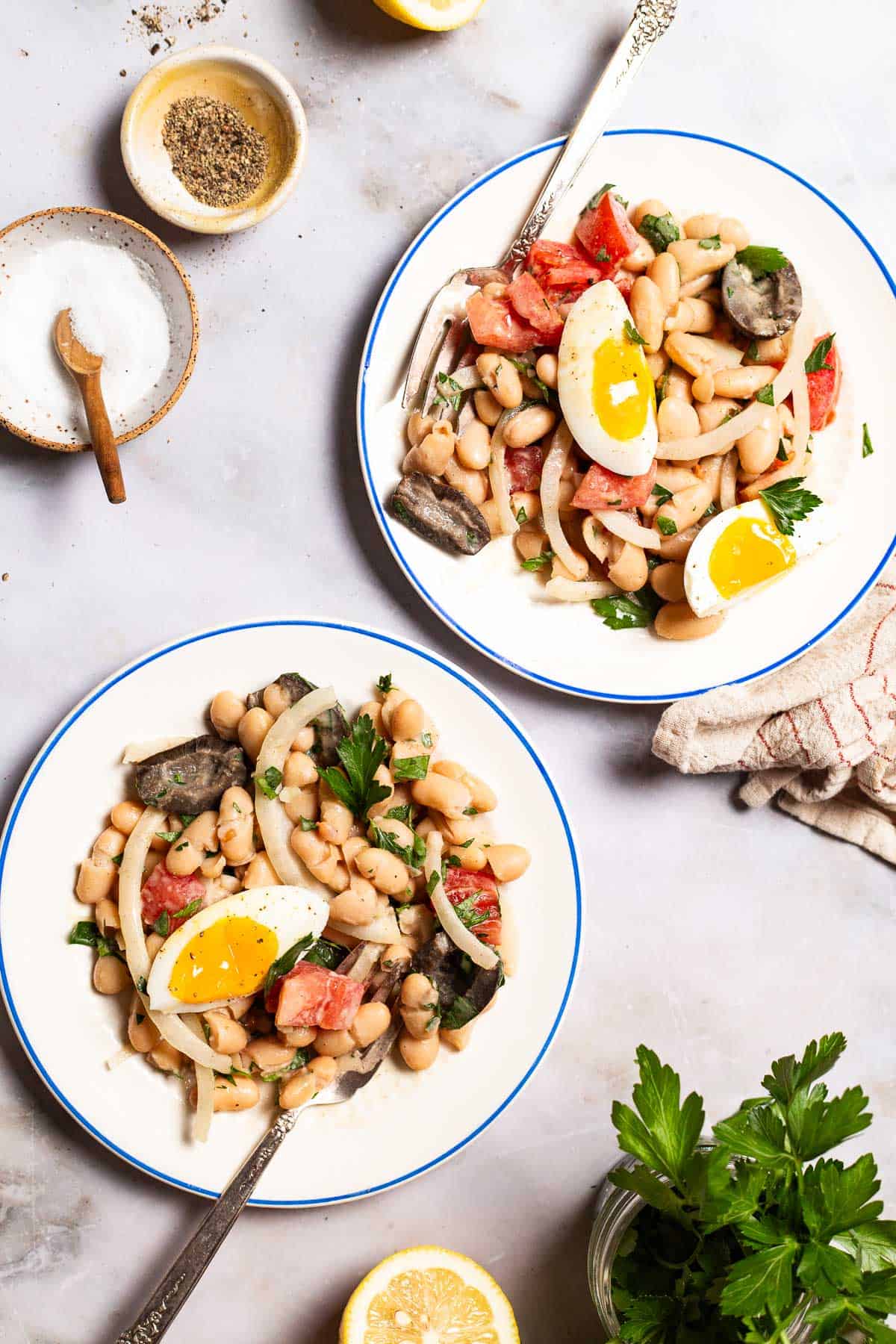 An overhead photo of two servings of cannellini bean salad on plates with forks. Next to these are small bowls of salt and pepper, a lemon half, some parsley and a cloth napkin.