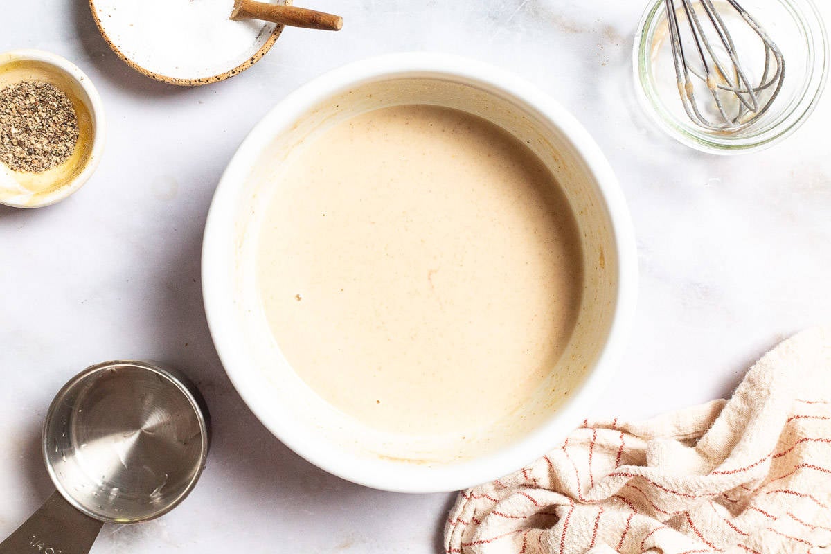 The tahini sauce for the cannellini bean salad in a bowl surrounded by small bowls of salt and pepper, a measuring cup, a whisk in a bowl and a cloth napkin.