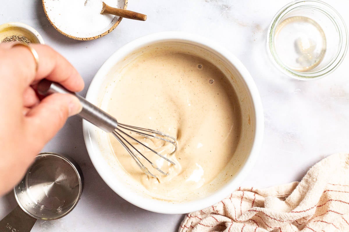 Tahini sauce being mixed in a bowl with a whisk. Next to this is a measuring cup, a cloth napkin, and small bowls of salt, pepper and water.