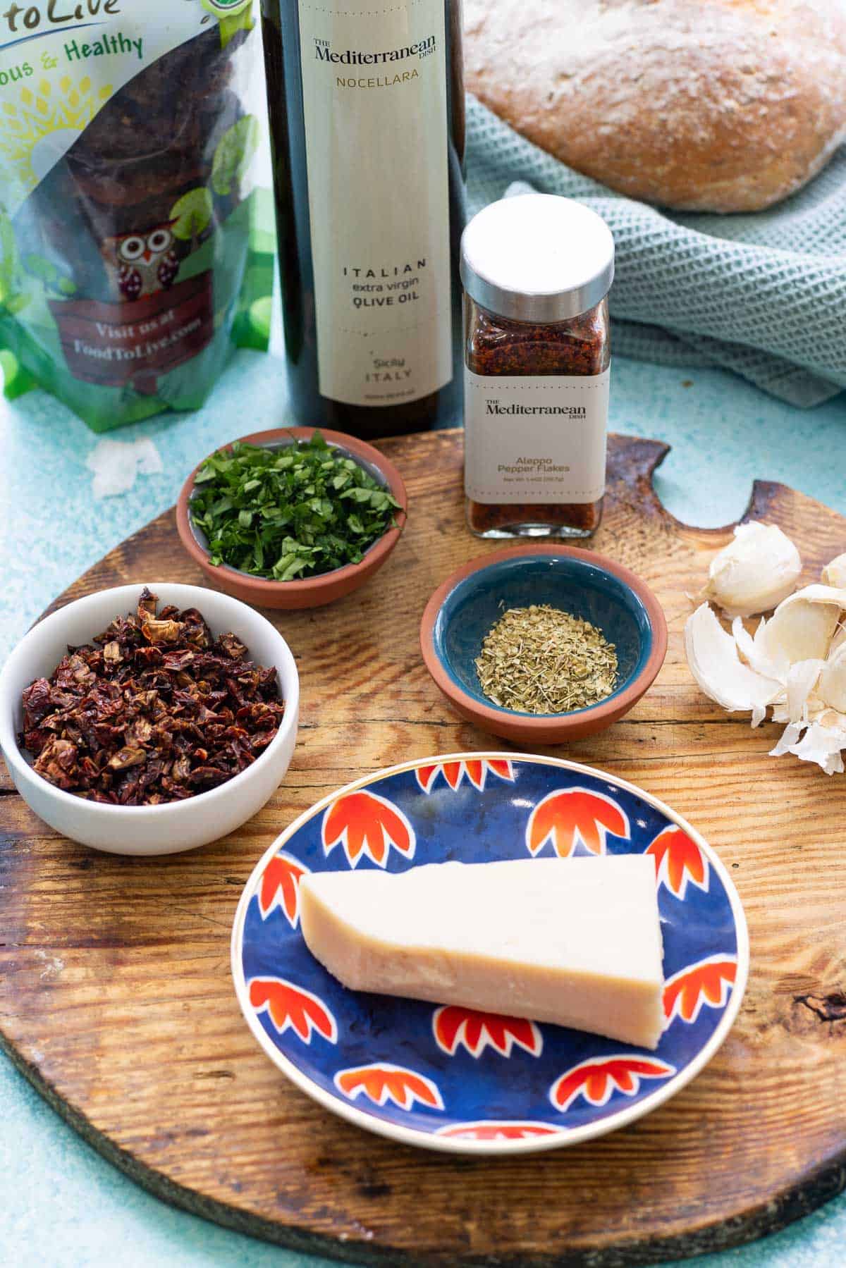 Ingredients for bread dipping oil including olive oil, sun dried tomatoes, Aleppo flakes, garlic, Italian seasoning, parsley, parmesan cheese and a loaf of crusty bread.