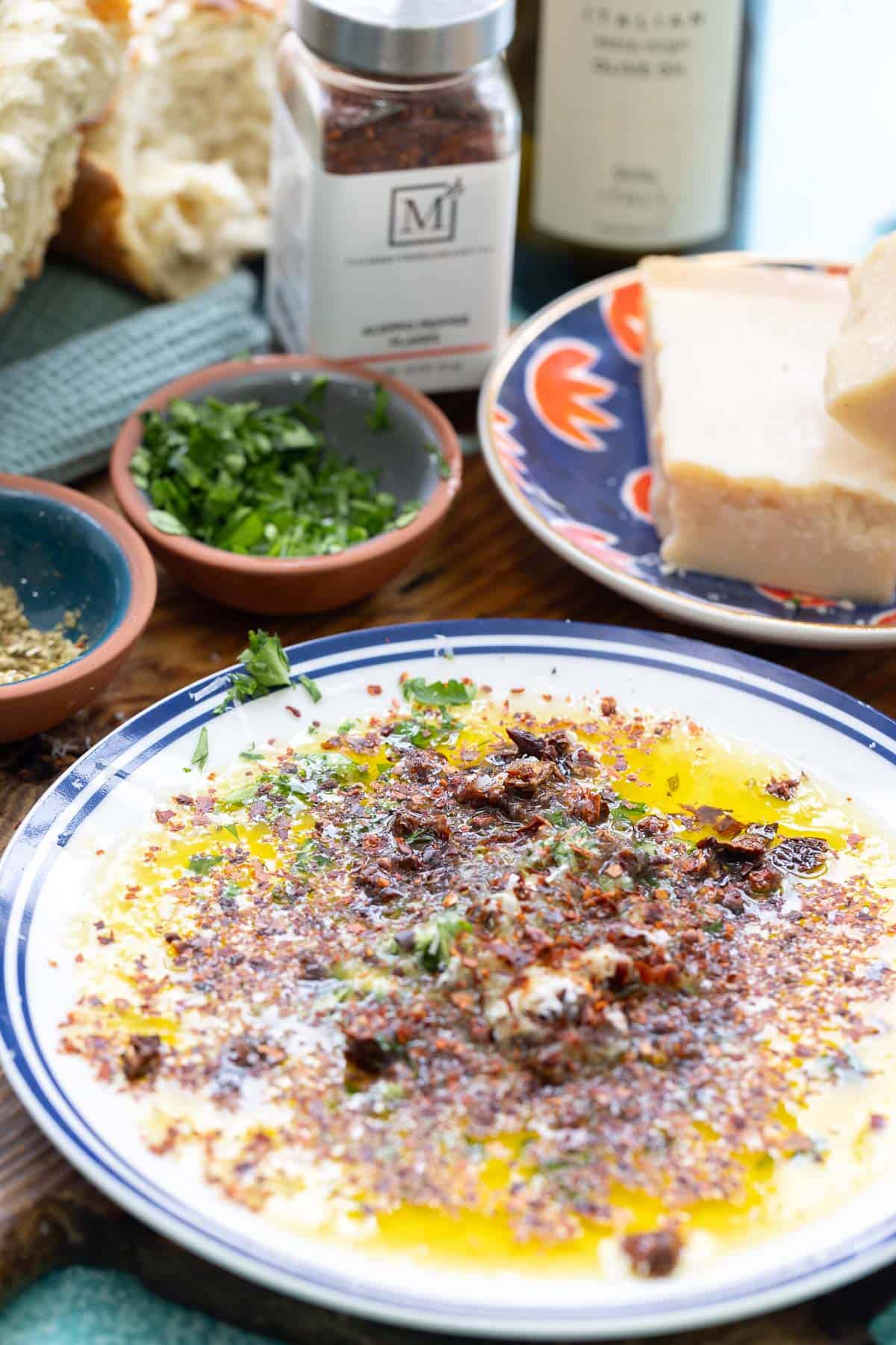 Bread dipping oil on a plate surrounded by small bowls of Italian seasoning and parsley, a wedge of parmesan cheese on a plate, pieces of crusty bread, a bottle of Aleppo pepper and olive oil.