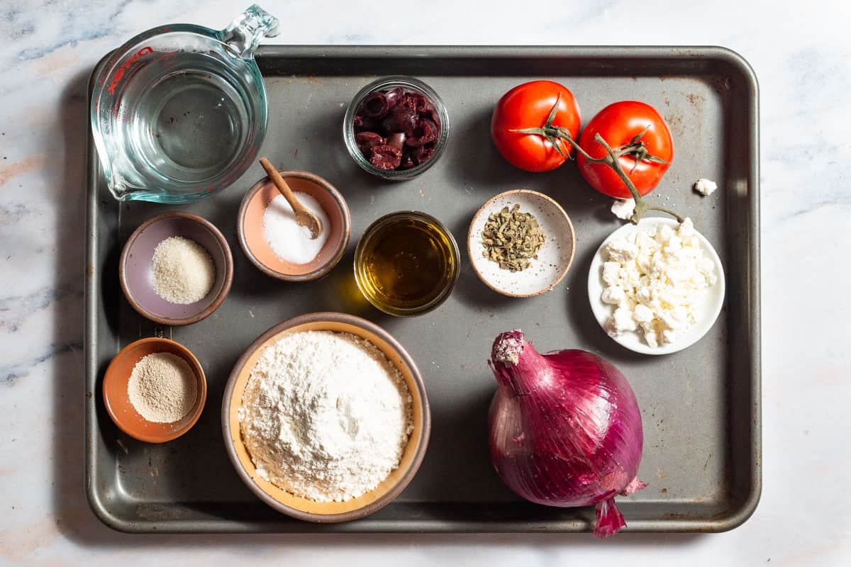Ingredients for Greek pizza including flour, yeast, superfine sugar, water, olive oil, salt, tomatoes, red onion, oregano, kalamata olives and feta.