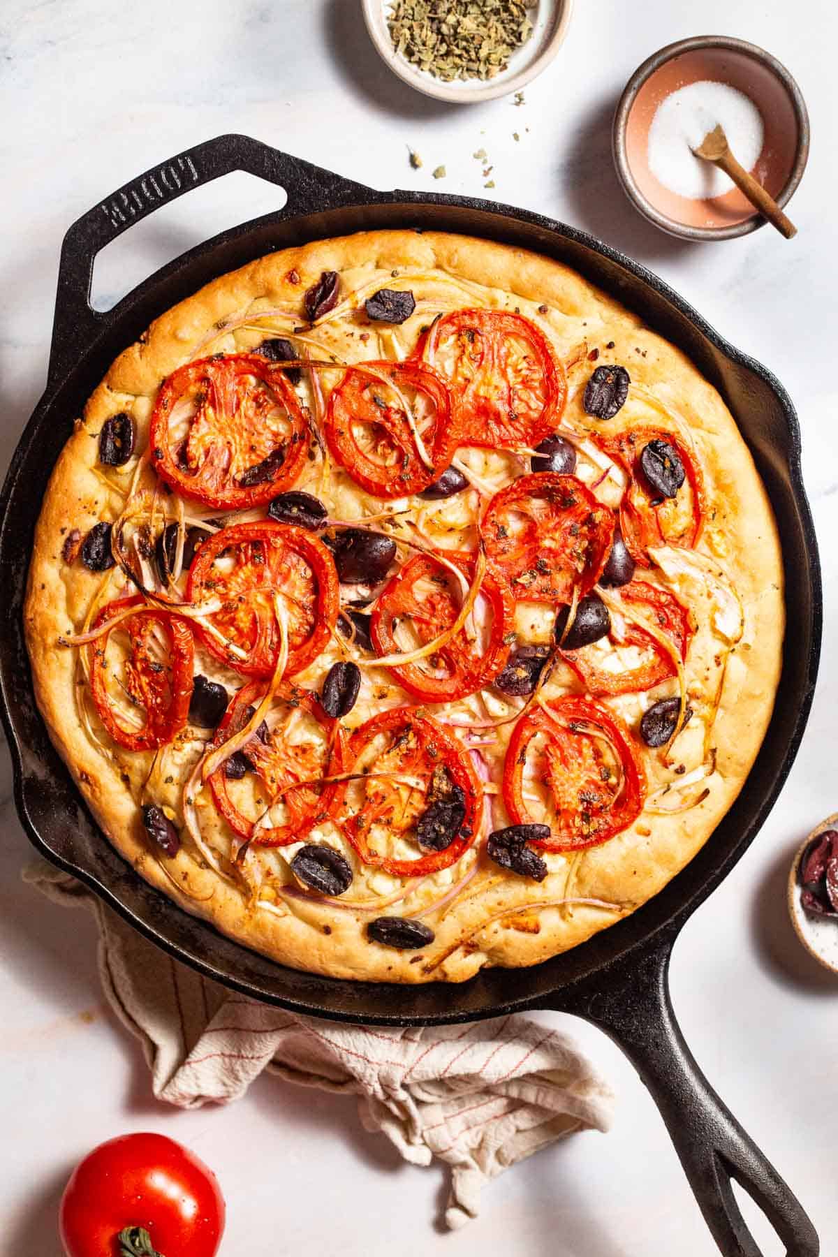 An overhead photo of greek pizza in a cast iron skillet. Surrounding this is a tomato, a kitchen towel, and small bowls of oregano and salt.
