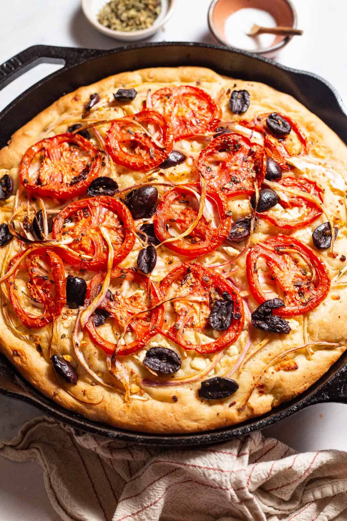 Greek pizza in a cast iron skillet next to a kitchen towel, and small bowls of oregano and salt.