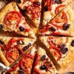 An entire sliced Greek pizza on a wooden cutting board next to bowls of oregano and kalamata olives.