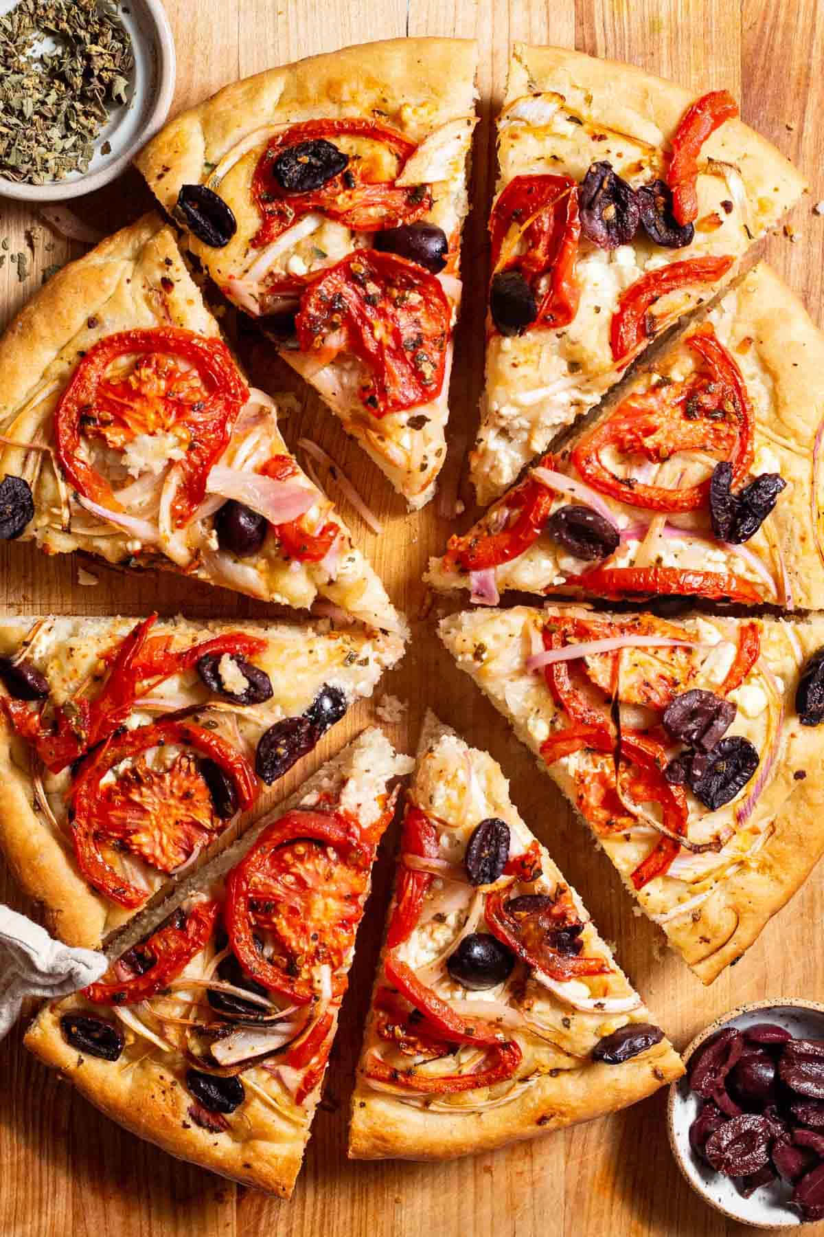 An entire sliced Greek pizza on a wooden cutting board next to bowls of oregano and kalamata olives.