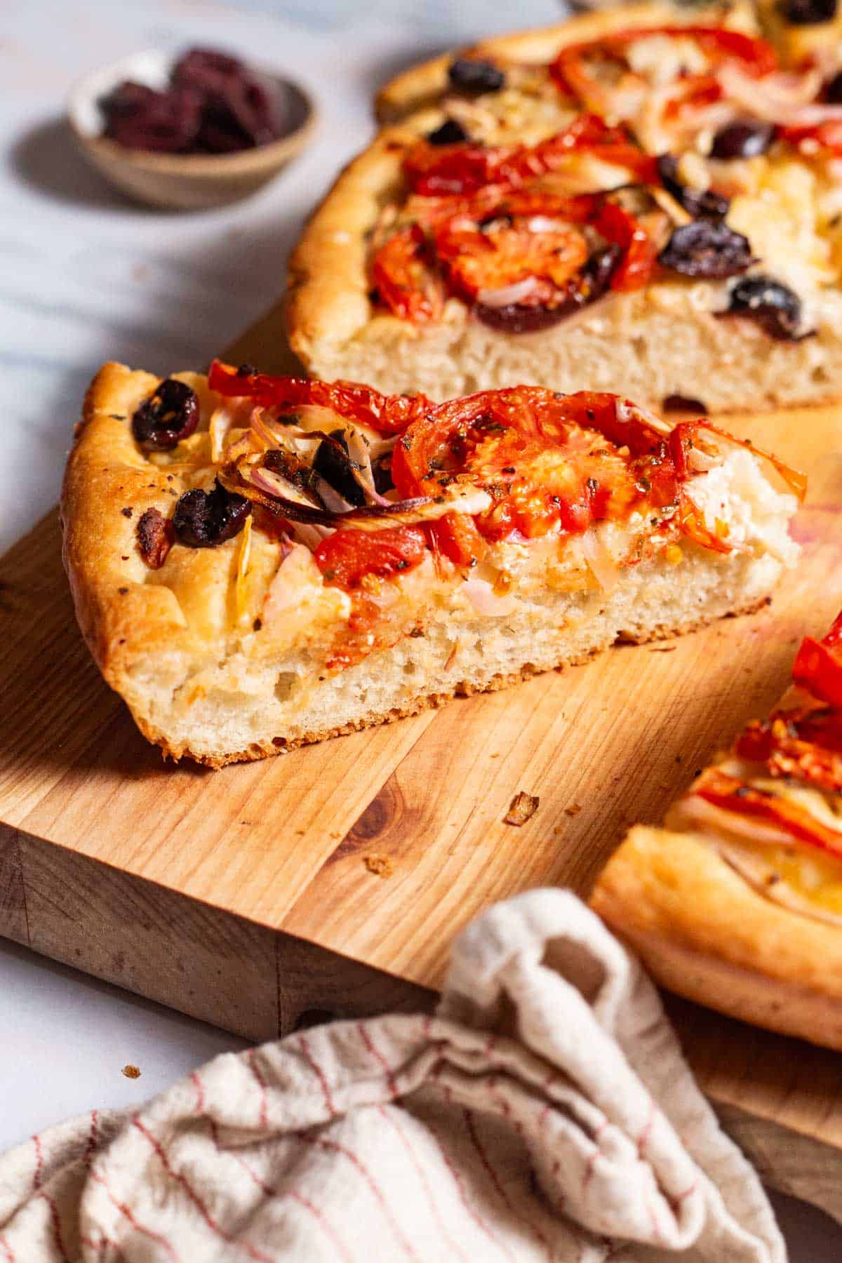 A close up photo of slices of Greek pizza on a cutting board. Next to these is a kitchen towel and a small bowl of kalamata olives.