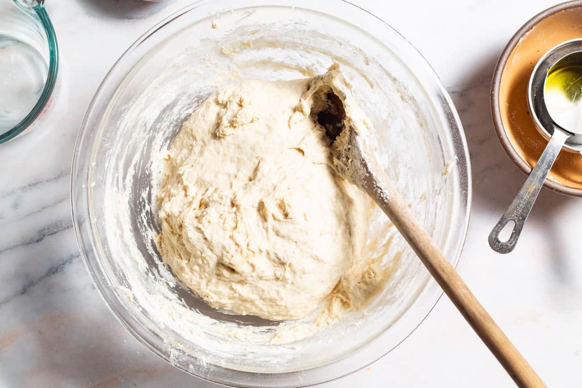 Pizza dough in a mixing bowl with a wooden spoon next to a measuring cup and a stack of bowls with another measuring cup.