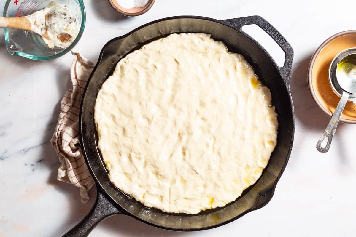 Pizza dough pressed into the bottom of a cast iron skillet. Next to this is a kitchen towel, a measuring cup with a wooden spoon in it, a small bowl of salt, and a stack of bowls with another measuring cup.