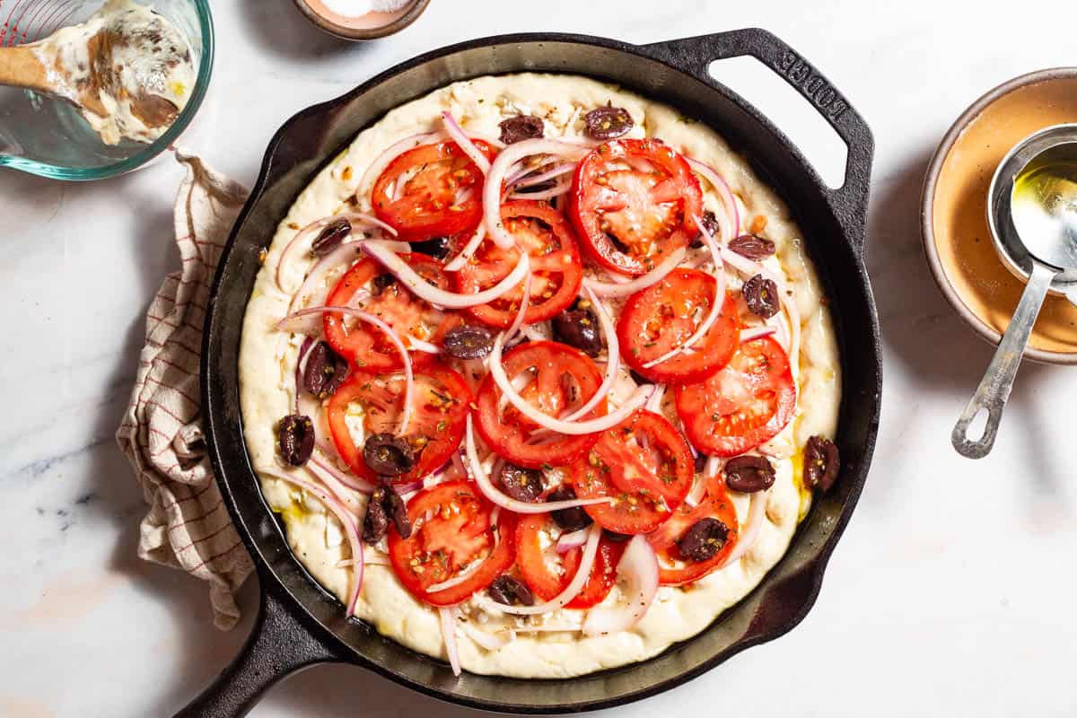 Greek pizza in a cast iron skillet before being baked. Next to this is a kitchen towel, a measuring cup with a wooden spoon, a small bowl of salt and a stack of bowls with another measuring cup.