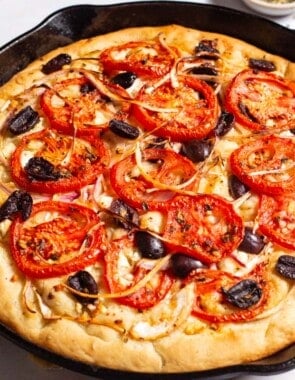 A close up of greek pizza in a cast iron skillet next to small bowls of kalamata olives and salt, and a kitchen towel.