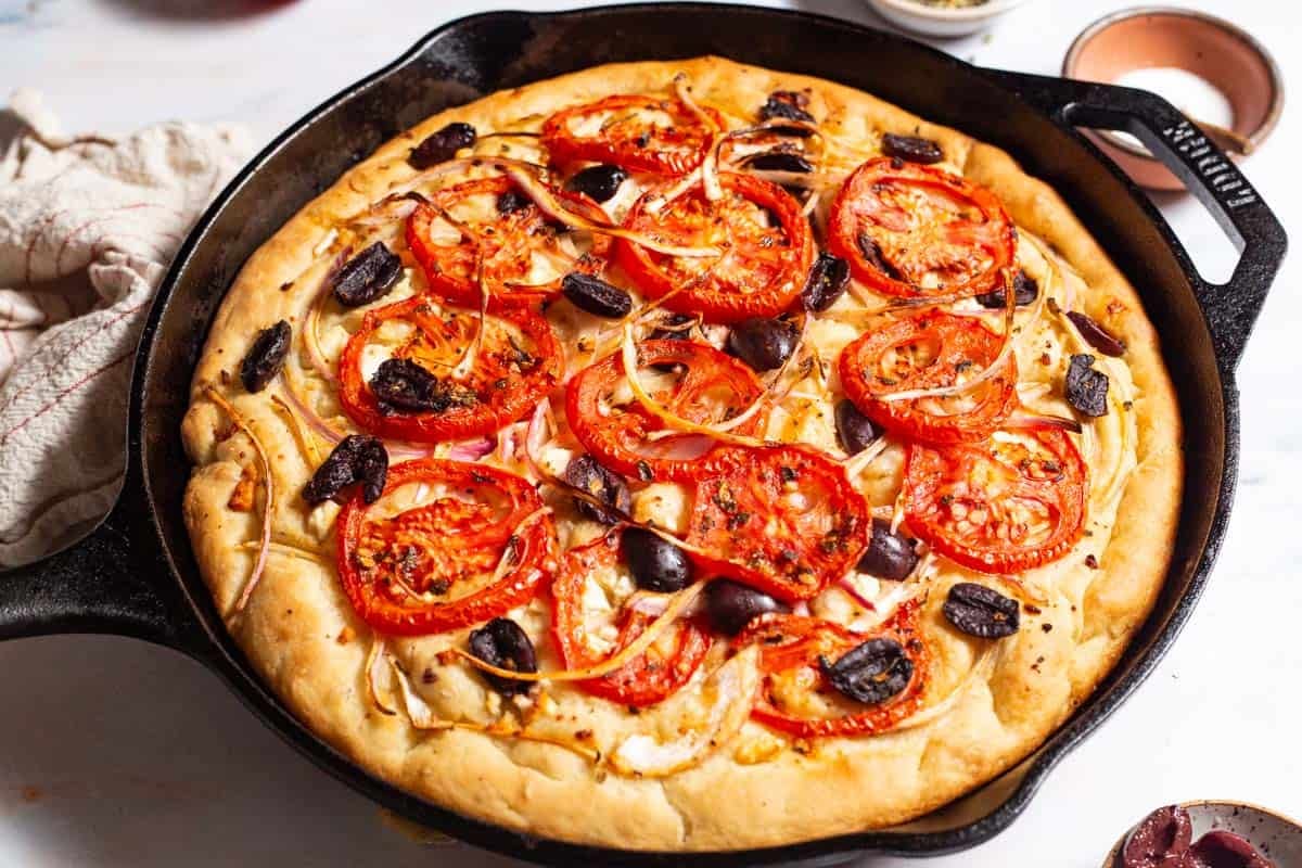 A close up of greek pizza in a cast iron skillet next to small bowls of kalamata olives and salt, and a kitchen towel.