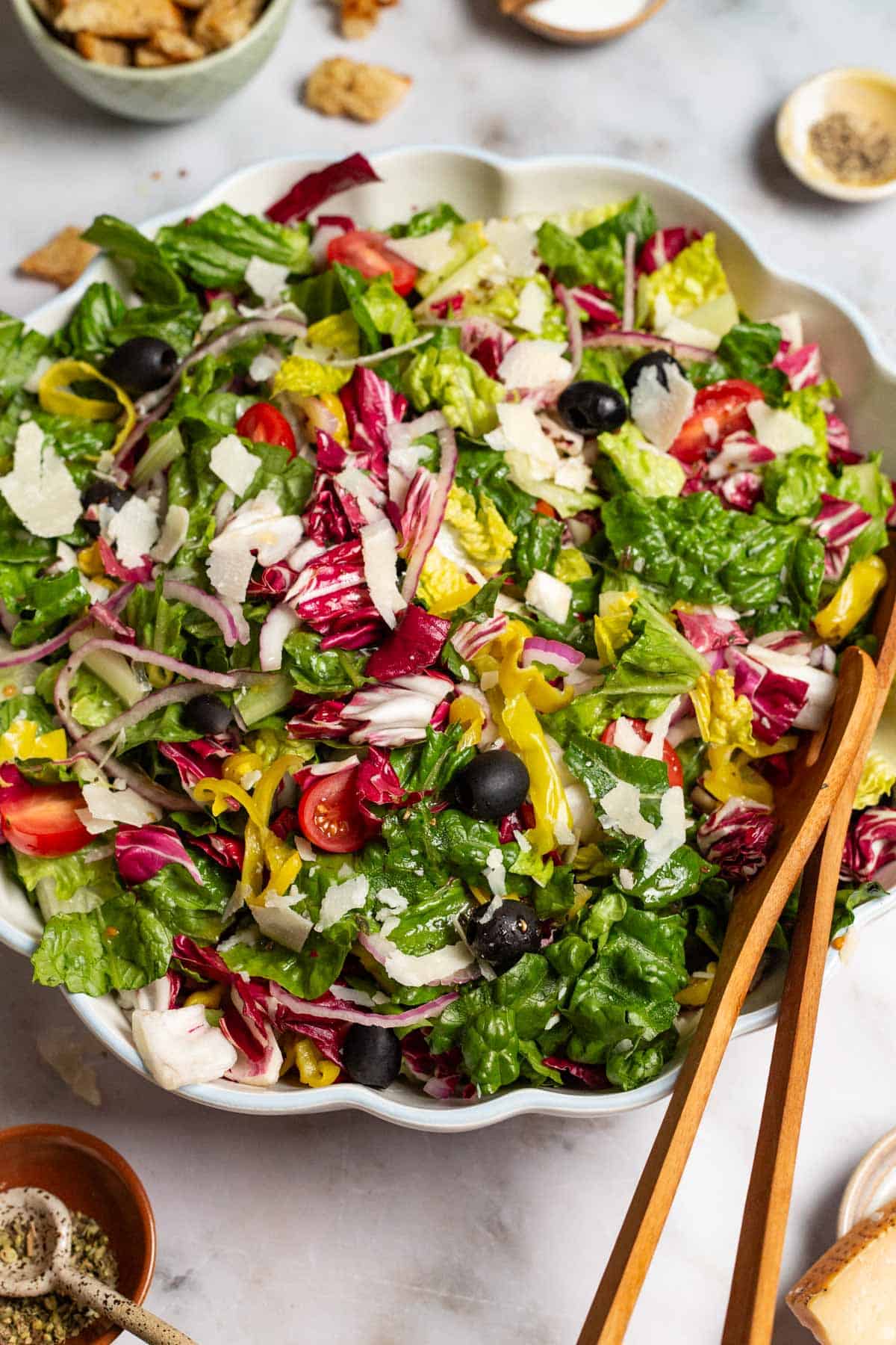 Italian salad in a serving bowl with wooden serving utensils. Next to this are bowls of croutons, salt, pepper and Italian seasoning.