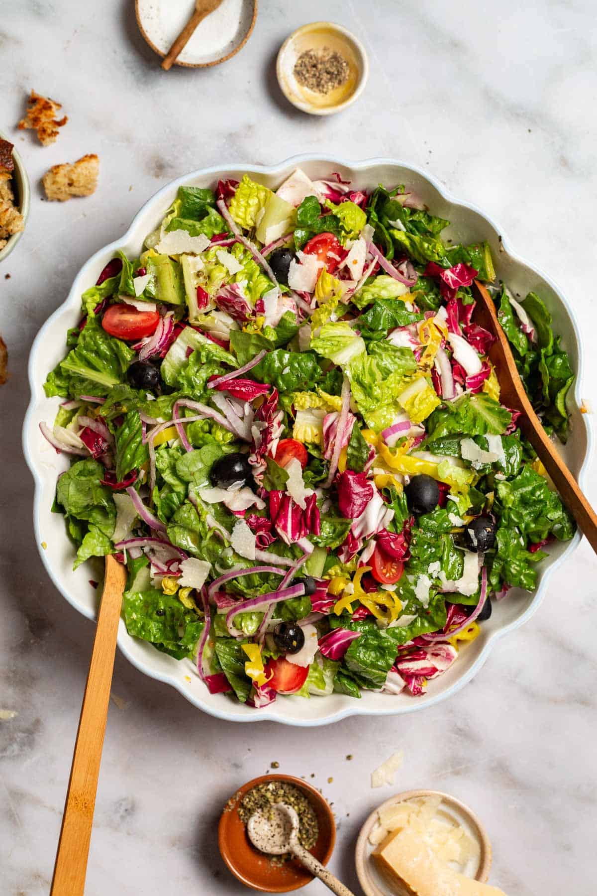 Italian salad in a serving bowl with wooden serving utensils. Next to this are bowls of salt, pepper, parmesan cheese and Italian seasoning.