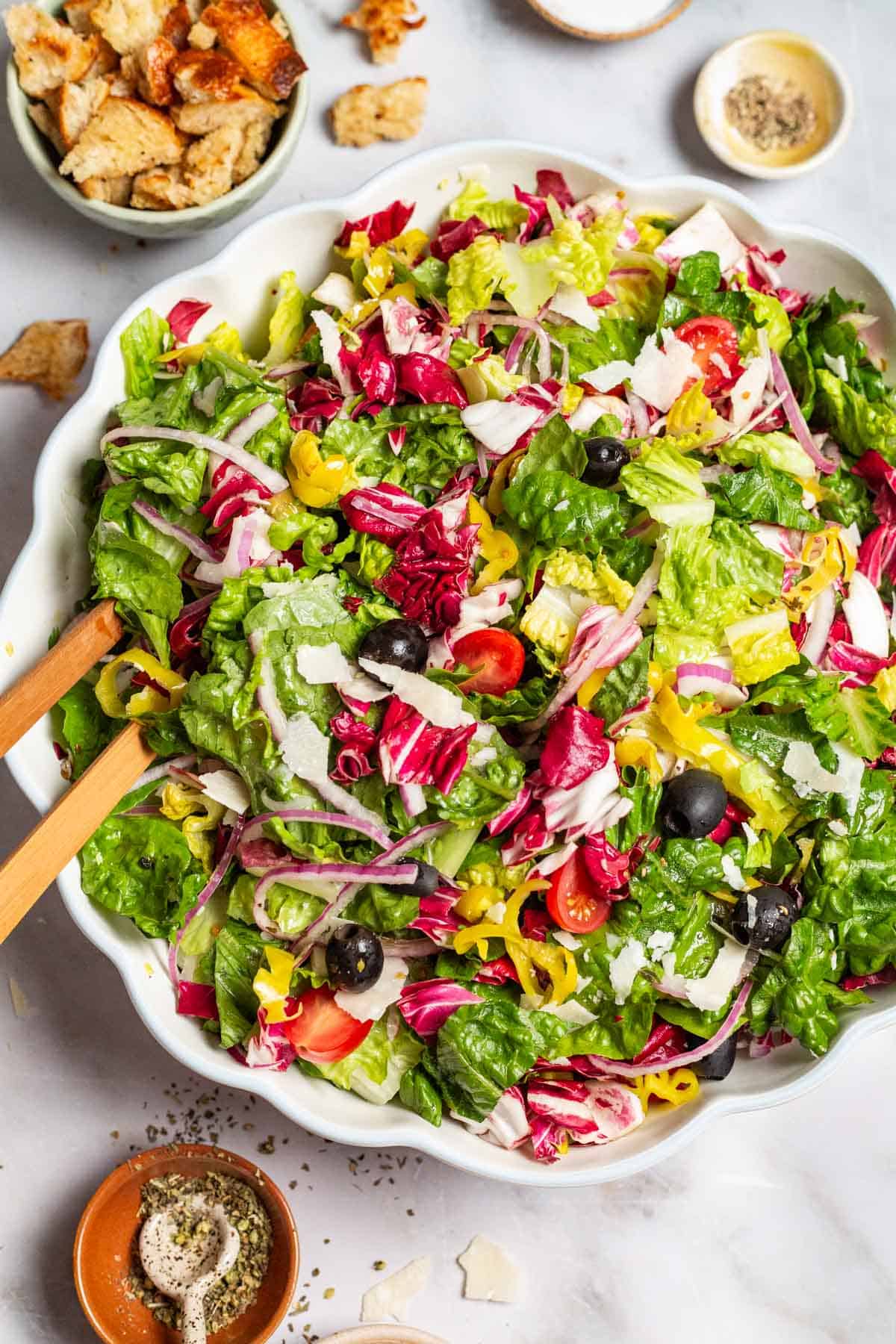 Italian salad in a serving bowl with wooden serving utensils. Next to this are bowls of croutons, salt, pepper and Italian seasoning.