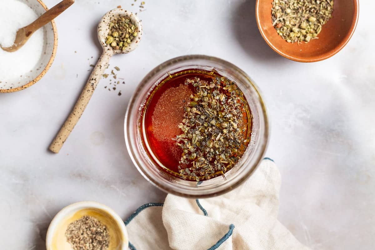 Italian seasoning added to a jar with red wine vinegar. Surrounding this are small bowls of salt, pepper and Italian seasoning, a spoon with Italian seasoning, and a cloth napkin.