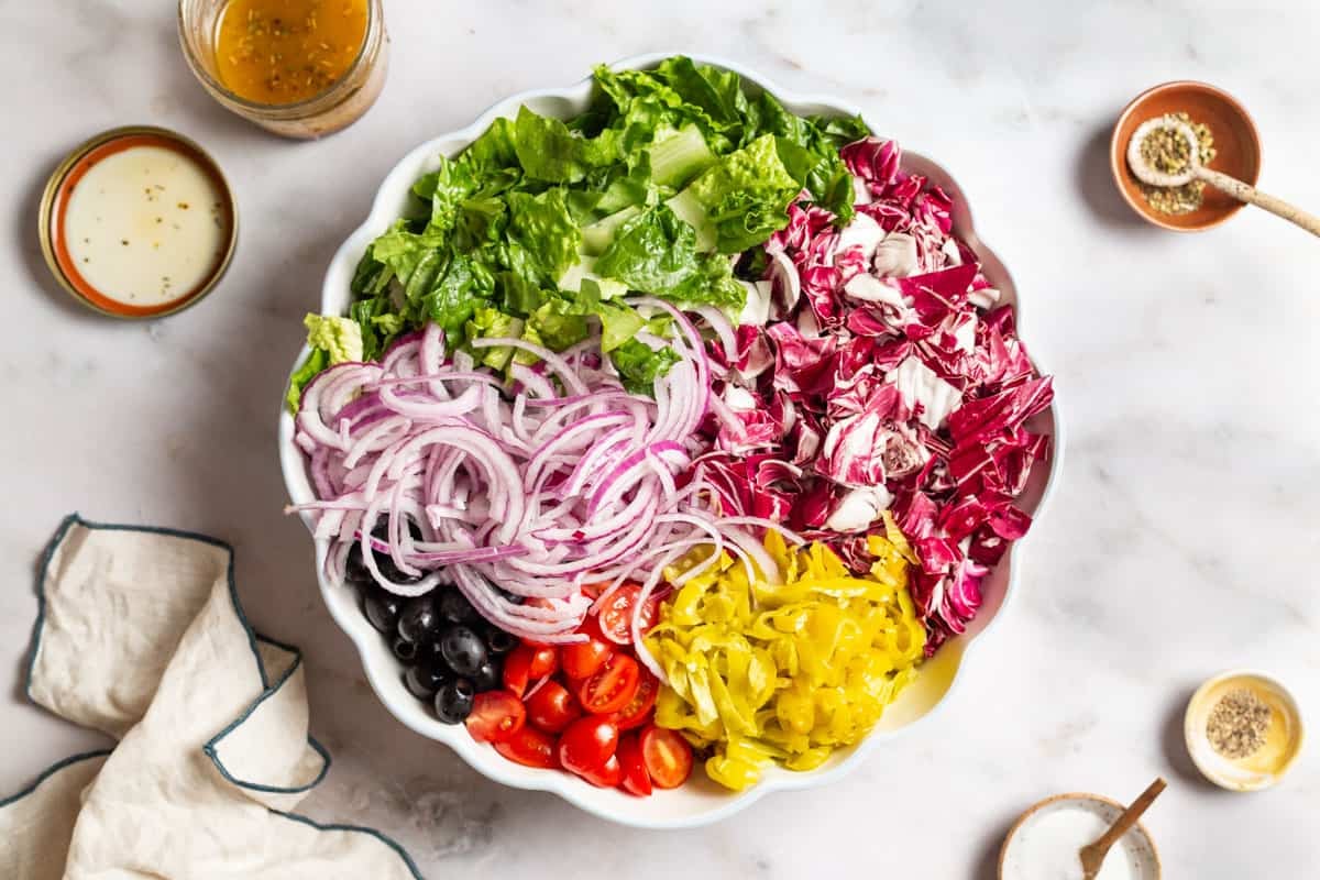 The ingredients for the Italian salad in a large serving bowl just before being mixed together. Surrounding this is a jar of the salad dressing and the lid, a cloth napkin, and small bowls of salt, pepper, and Italian seasoning.