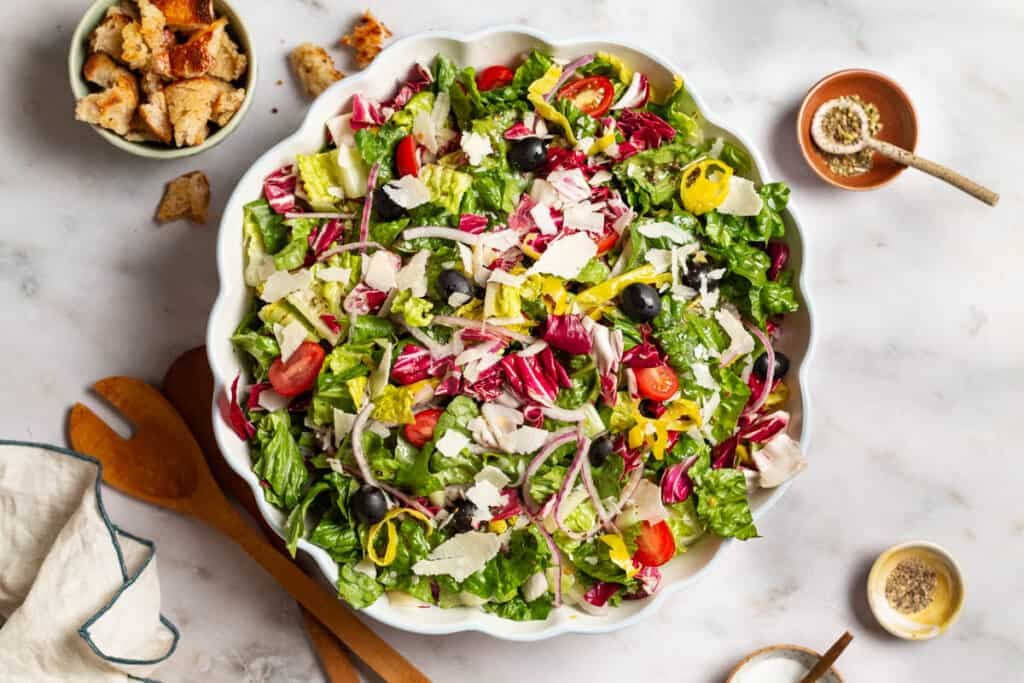 Italian salad in a serving bowl. Next to this are wooden serving utensils, a cloth napkin, and bowls of croutons, salt, pepper and Italian seasoning.