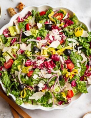 Italian salad in a serving bowl. Next to this are wooden serving utensils, a cloth napkin, and bowls of croutons, salt, pepper and Italian seasoning.