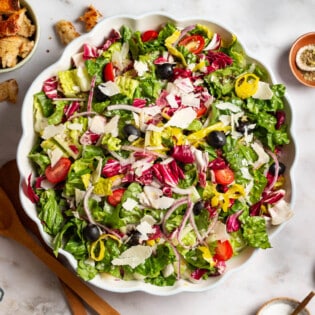 Italian salad in a serving bowl. Next to this are wooden serving utensils, a cloth napkin, and bowls of croutons, salt, pepper and Italian seasoning.