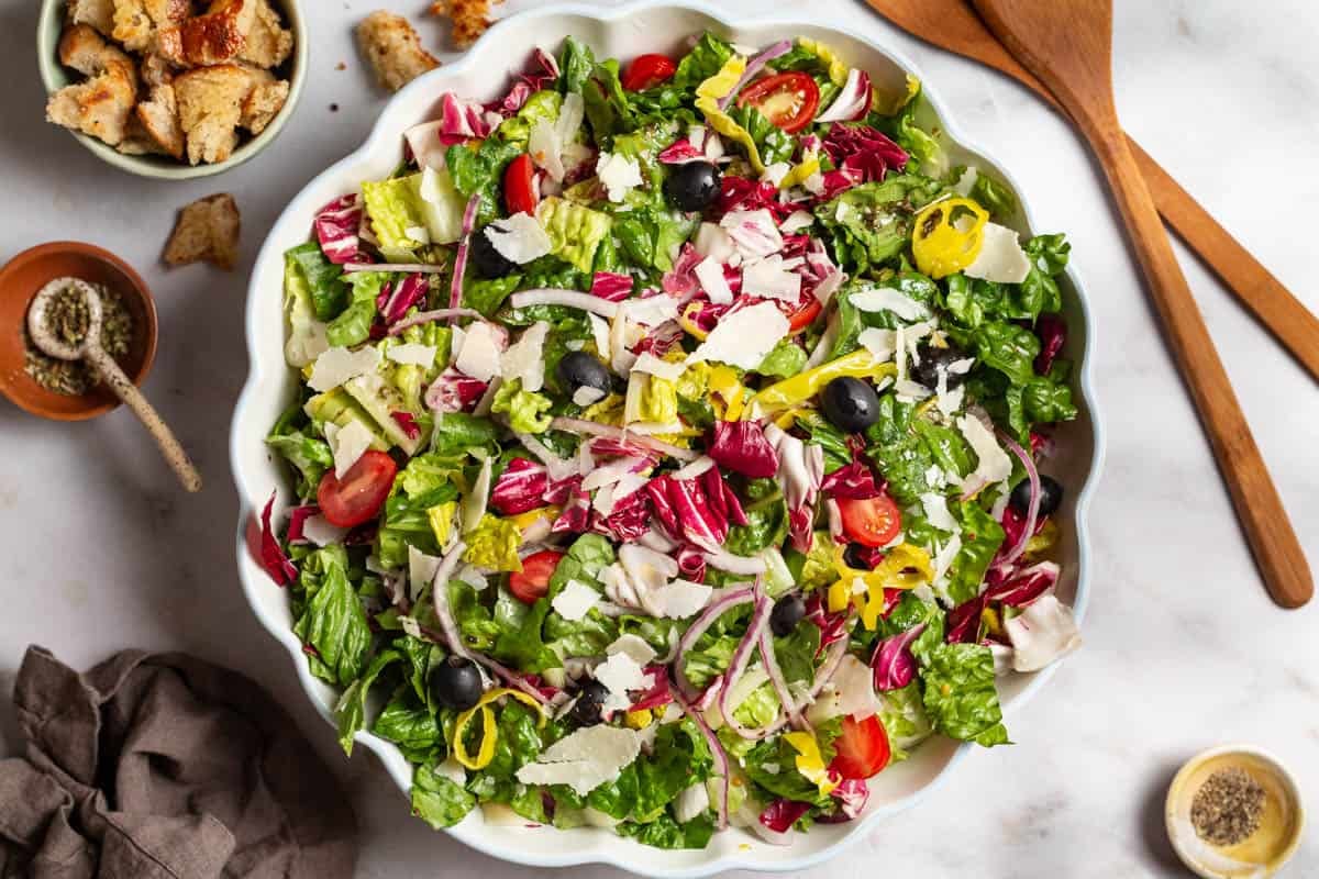 Italian salad in a serving bowl. Next to this are wooden serving utensils, a cloth napkin, and bowls of croutons, pepper and Italian seasoning.