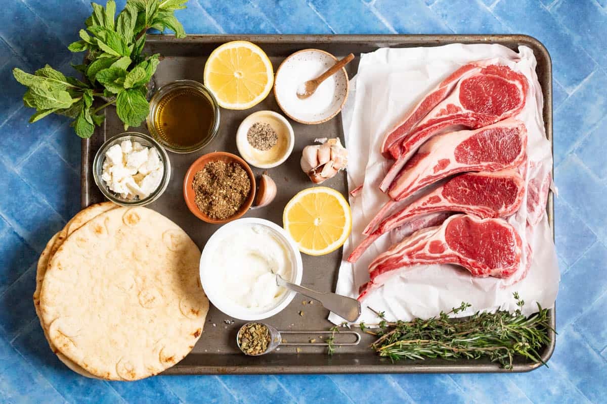 Ingredients for pan seared lamb chops including lamb chops, salt, pepper, oregano, olive oil, garlic, thyme, rosemary, lemons, pita bread, za'atar, feta, greek yogurt and mint.