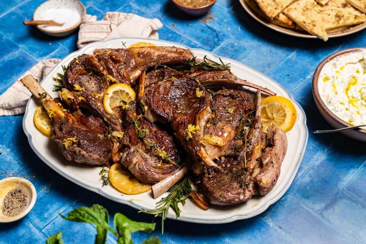 Pan seared lamb chops on a serving platter. This is surrounded by a plate of herbed flatbread, a bowl of yogurt sauce, small bowls of za'atar, salt and pepper, a kitchen towel and some mint.