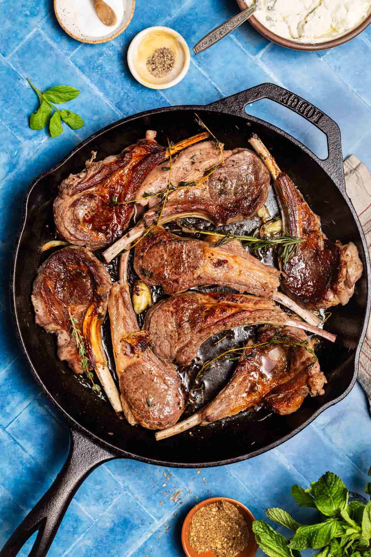 An overhead photo of pan seared lamb chops in a cast iron skillet surrounded by small bowls of salt, pepper and za'atar, some mint, and a bowl of the yogurt sauce.