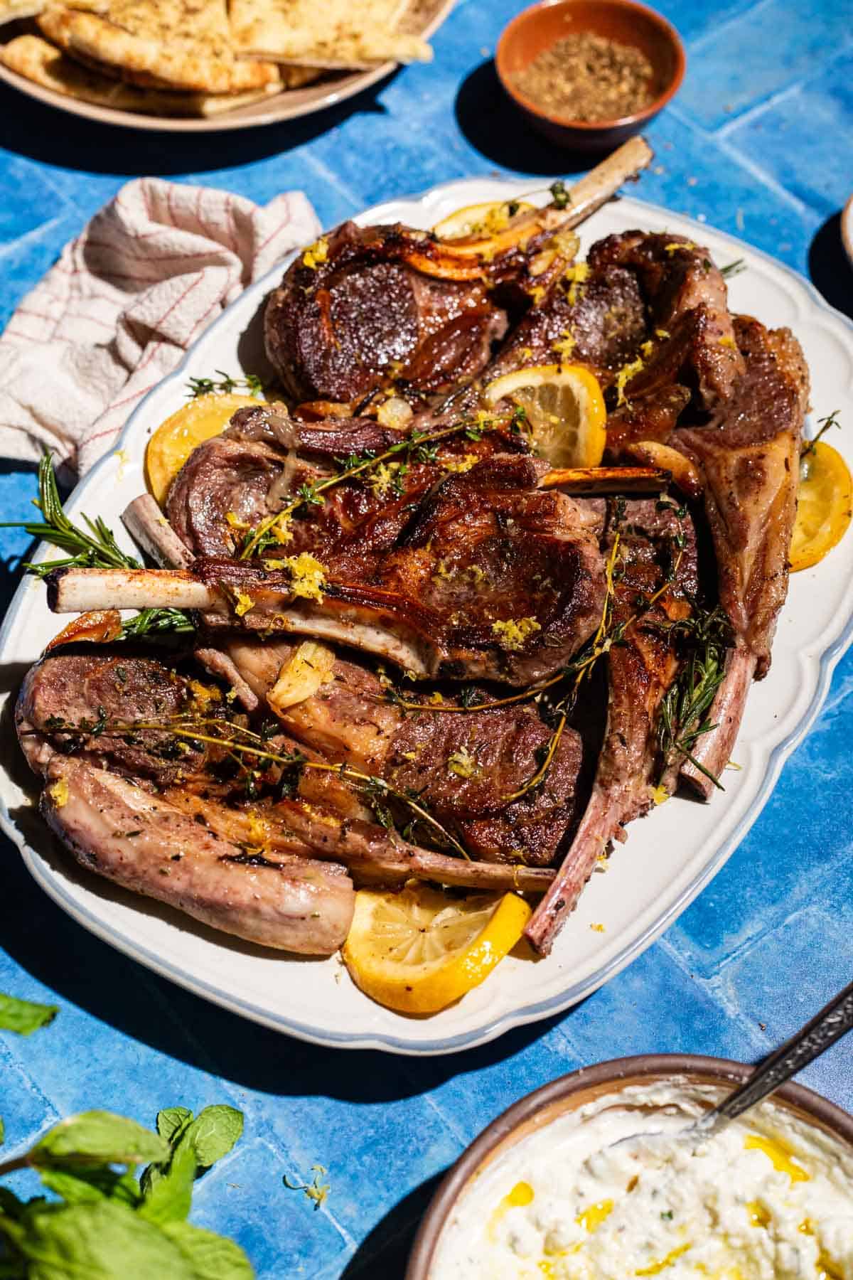 Pan seared lamb chops on a serving platter. This is surrounded by a plate of herbed flatbread, a bowl of yogurt sauce, a small bowl of za'atar, a kitchen towel and some mint.