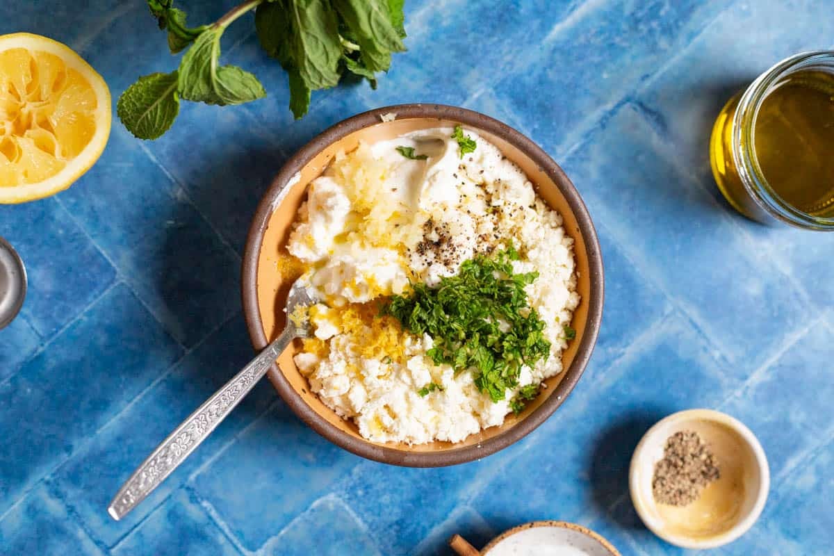 The ingredients for the yogurt sauce in a bowel just before being mixed together with a spoon. Next to this is a juiced lemon half, some mint, olive oil and small bowls of salt and pepper.