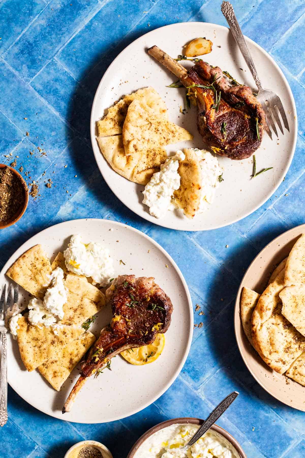 An overhead photo of two plates, each with a pan seared lamb chop, some seasoned flatbread, yogurt sauce and a fork. Next to this is a plate with more of the flatbread and bowls of za'atar and yogurt sauce.