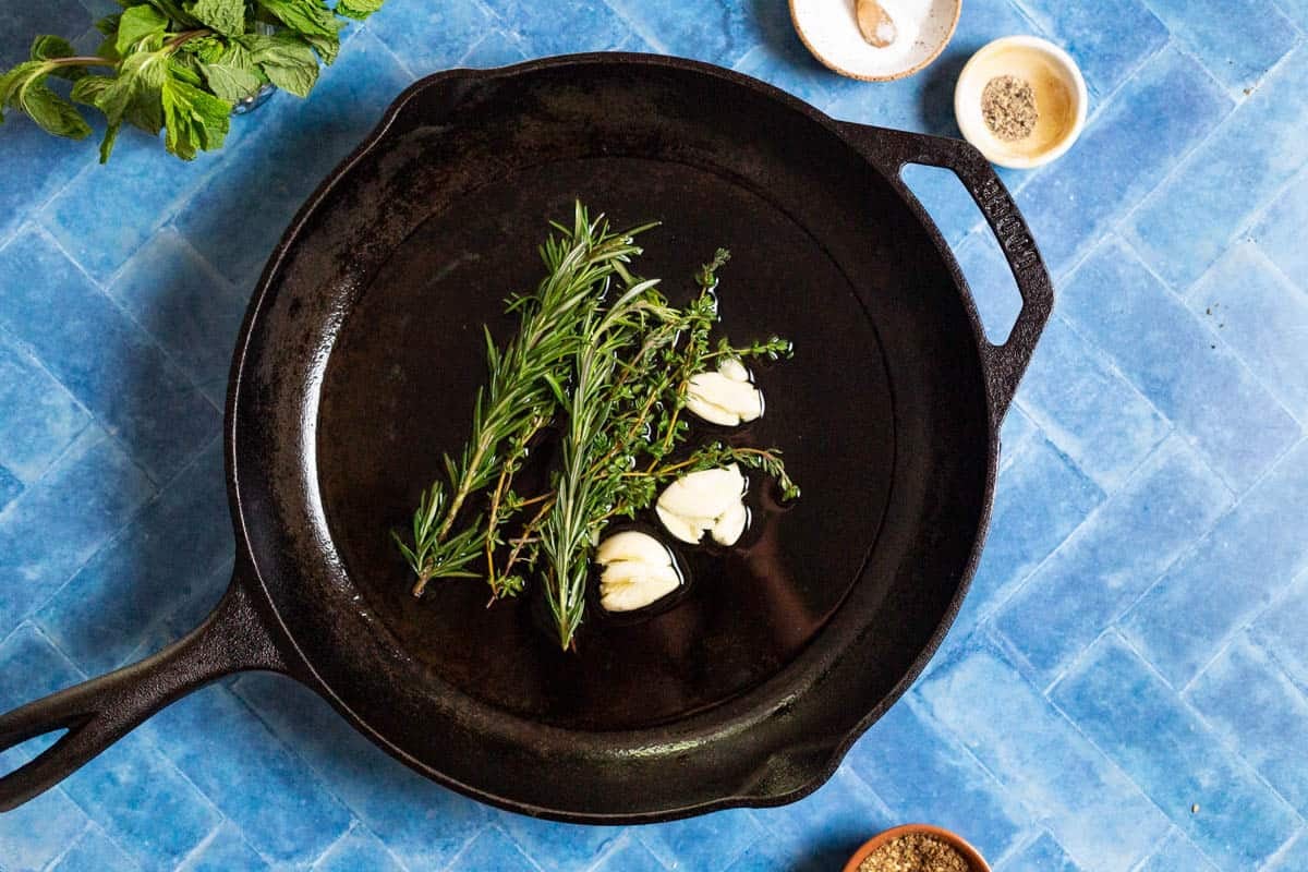 garlic, thyme and rosemary cooking in olive oil in a cast iron skillet. Surrounding this is some mint, and small bowls of salt, pepper and za'atar.