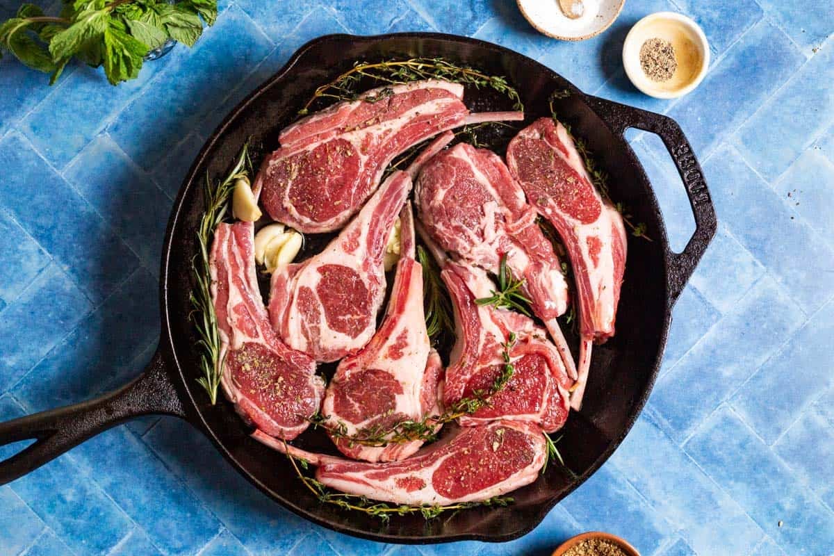 Uncooked lamb chops in a cast iron skillet with the garlic, rosemary and thyme. Surrounding this is some mint, and small bowls of salt, pepper and za'atar.