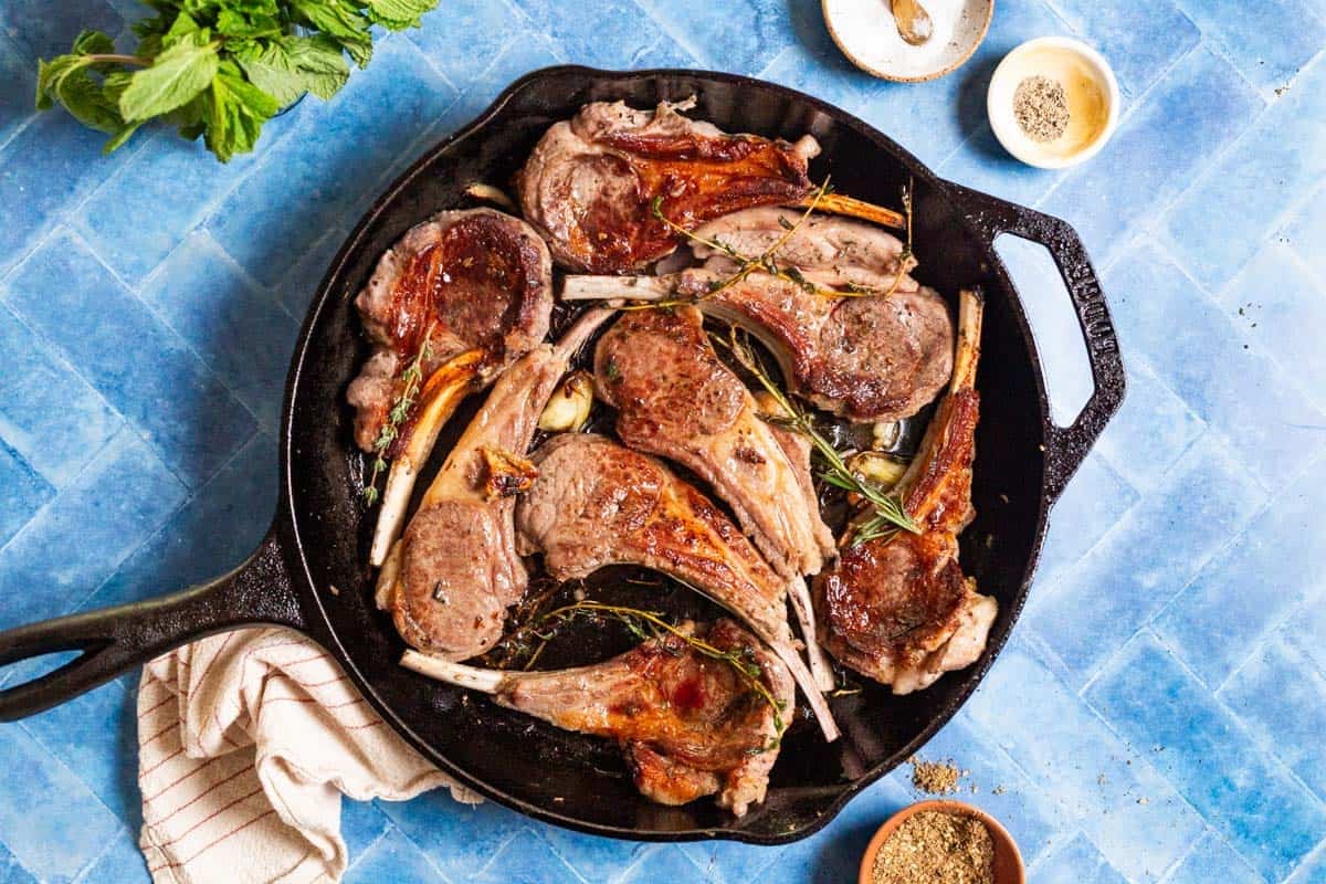 Pan seared lamb chops in a cast iron skillet surrounded by small bowls of salt, pepper and za'atar, some mint, and a kitchen towel.