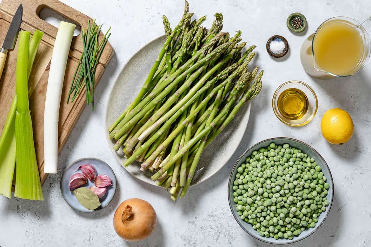 Ingredients for asparagus soup including asparagus, onion, garlic, olive oil, celery, a leek, vegetable broth, frozen peas, a bay leaf, salt, pepper, lemon juice, and chives.