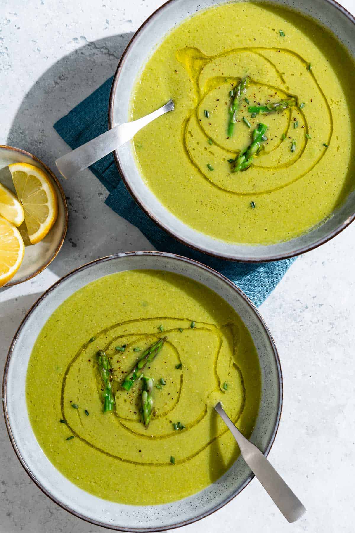 An overhead photo of two bowls of asparagus soup with spoons. Next to these is a small plate of lemon wedges.