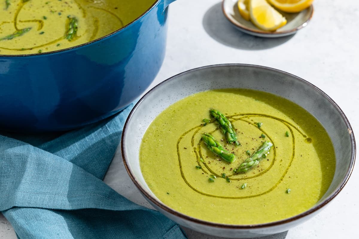 A close up of a bowl of asparagus soup. In the background is the pot of asparagus soup and a small plate of lemon wedges.