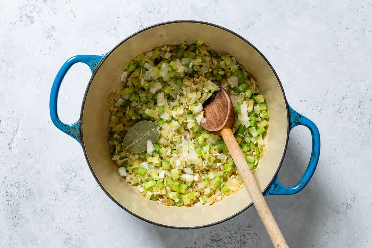 Chopped onion, celery, garlic, leeks and a bay leaf being sauteed in a large pot with a wooden spoon.