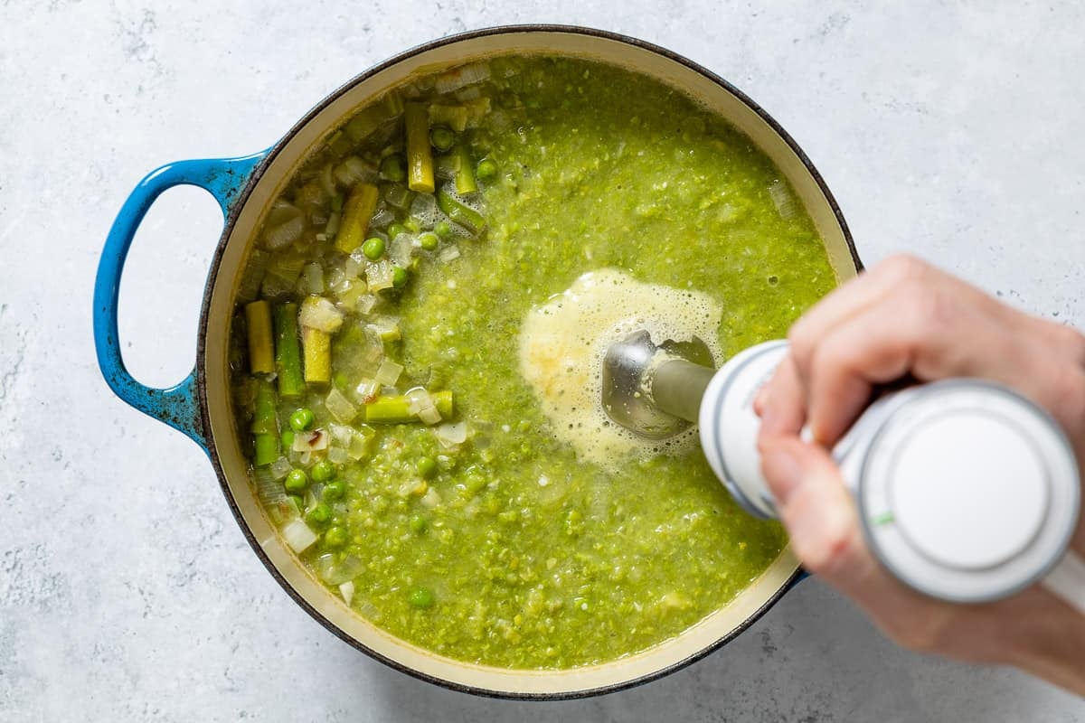 The cooked asparagus soup in a large pot being blended with an immersion blender.