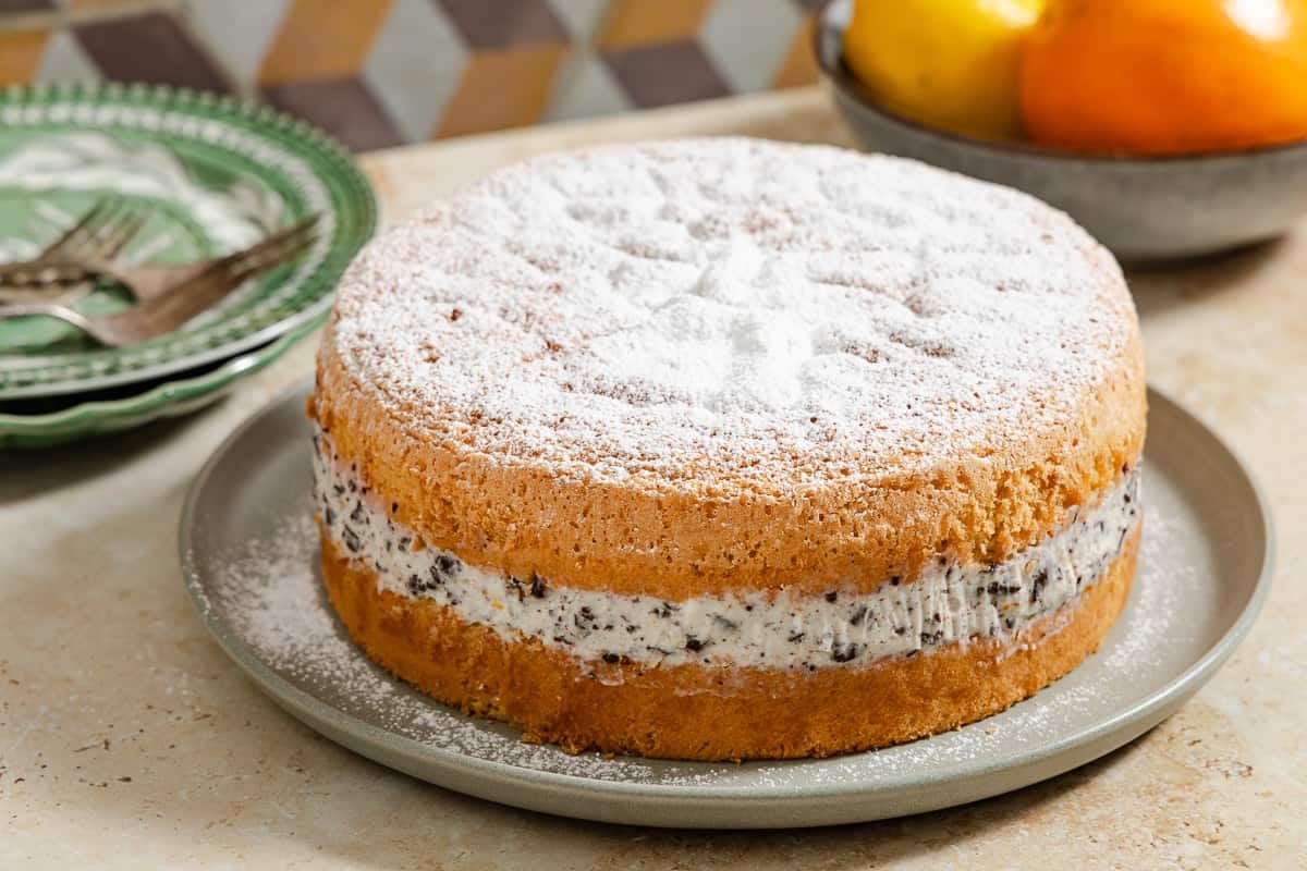 The easy cassata cake on a serving platter, with a stack of plates with forks and a bowl of lemons and oranges in the background.