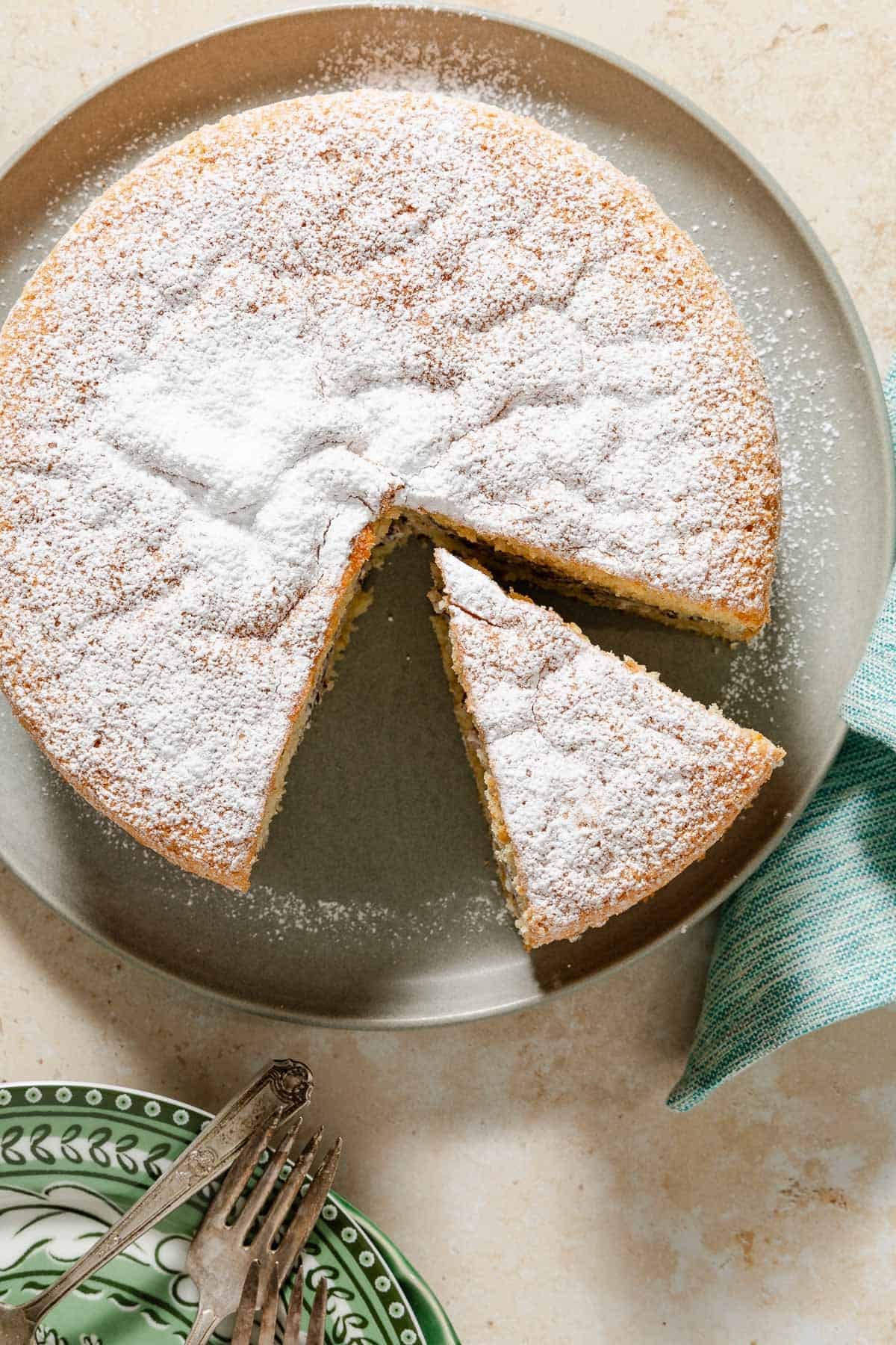 An overhead photo of the easy cassata cake on a platter with a slice cut and separated from it. Next to this is a stack of plates with forks.