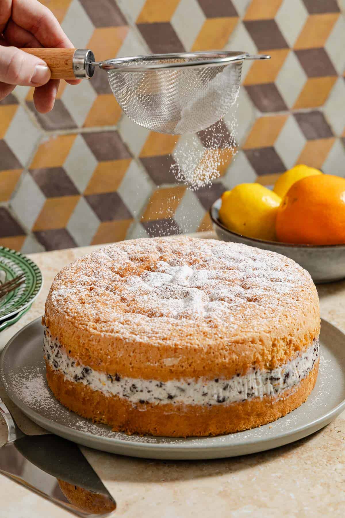 Powdered sugar being sprinkled on top of the easy cassata cake on a serving platter. Surrounding this is a stack of plates with forks and a bowl of citrus fruits.